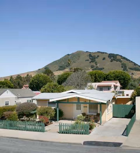 A small beige house with green trim, front yard, and a mountain backdrop.