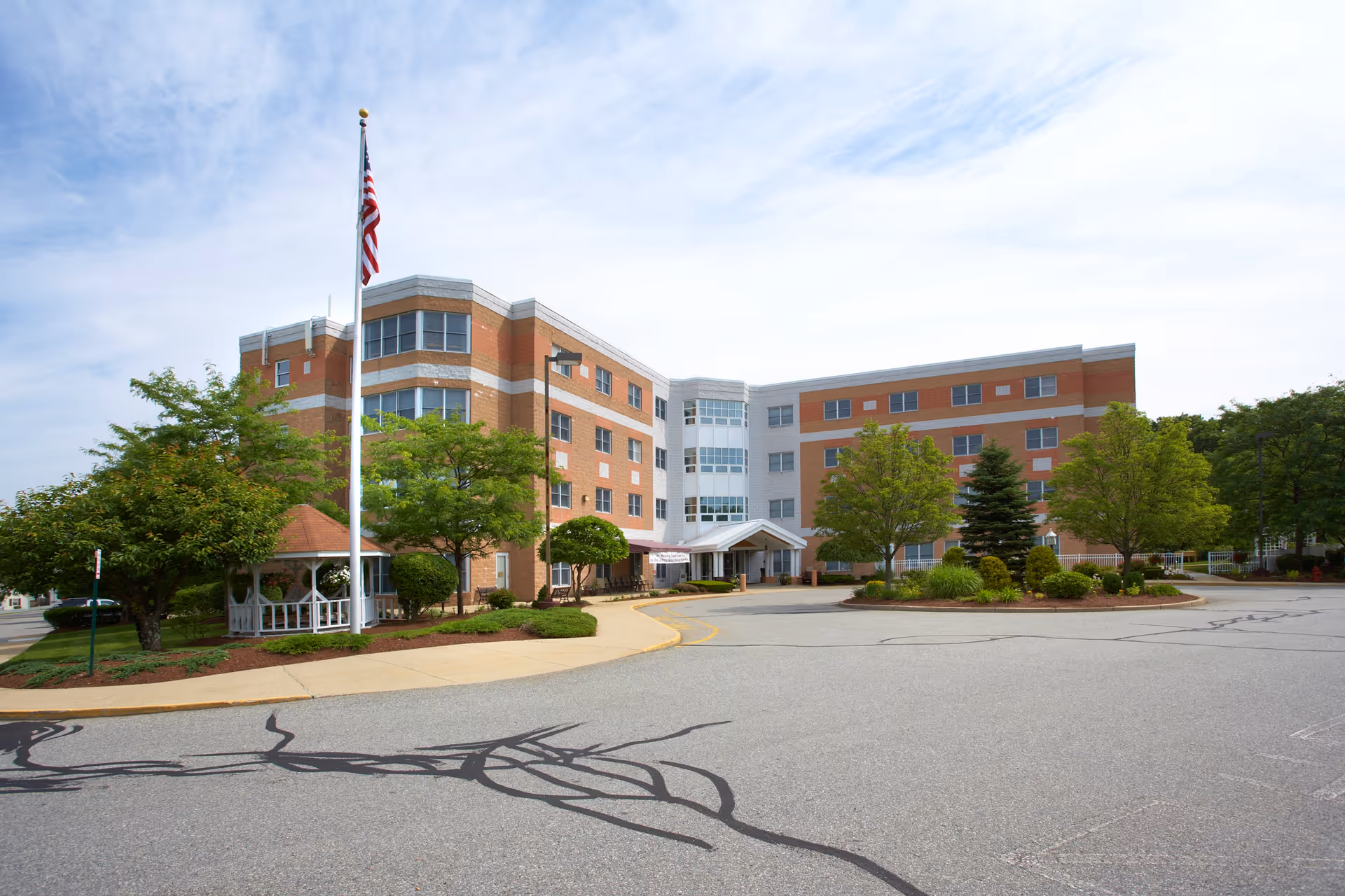 Front exterior of a multi-story brick health care facility with a flagpole, gazebo, trees, and a circular driveway.