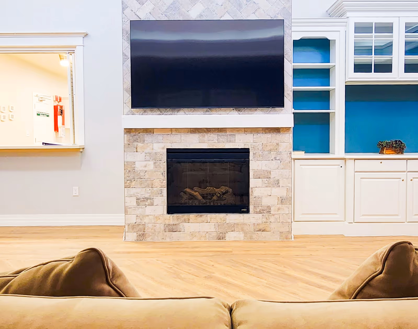 Interior view of a living room area in Barton House featuring a beige couch in the foreground, a stone fireplace with a mounted flat-screen TV above it, and built-in white cabinetry with blue backing on the right side. There is also a pass-through window to another room on the left side.