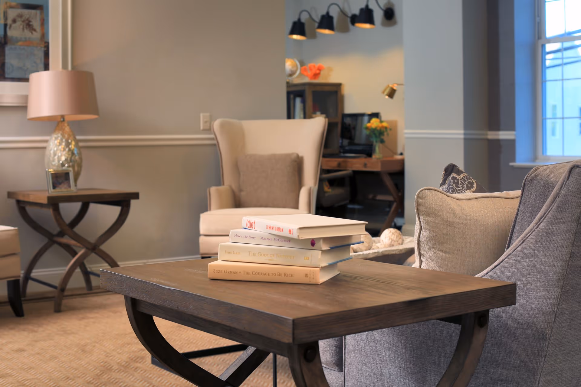 A cozy living room area with a wooden coffee table holding a stack of books, a beige armchair with a cushion, a side table with a lamp and a small framed photo, and a window letting in natural light. In the background, there is a desk with a computer and a vase with flowers.