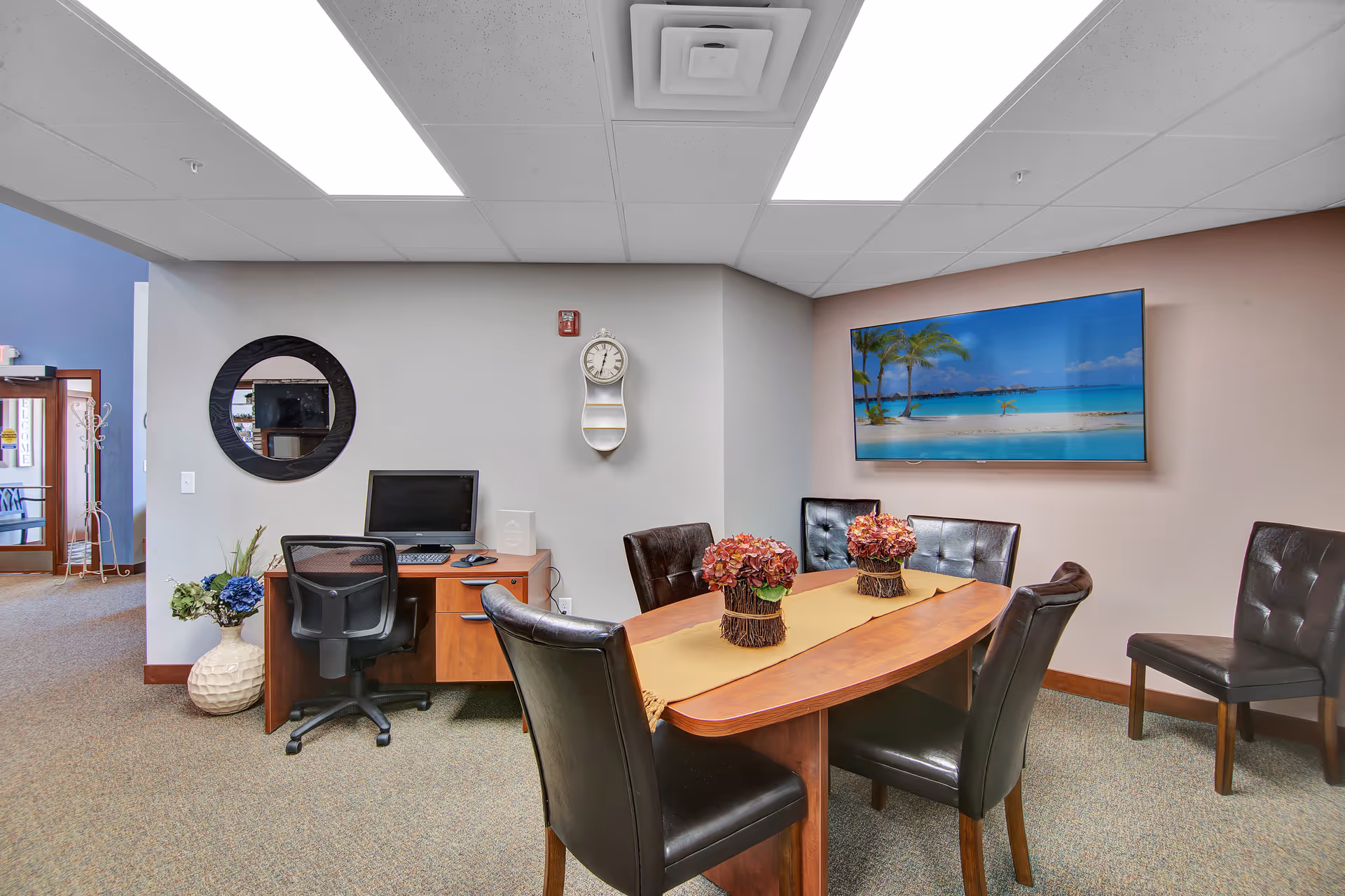 A senior living facility common area with a wooden table surrounded by six black leather chairs. Two floral centerpieces are on the table. A large flat-screen TV on the wall displays a beach scene with palm trees and blue water. To the left, there is a small wooden desk with a computer and a black office chair. A round mirror and a wall clock are mounted on the wall above the desk. A vase with flowers is on the floor next to the desk. The room has a carpeted floor and a drop ceiling with fluorescent lights.