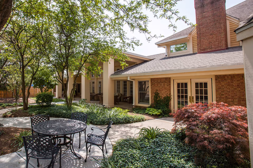 Sunny courtyard with a metal patio table and chairs amid trees and landscaping in front of a brick building with French doors.