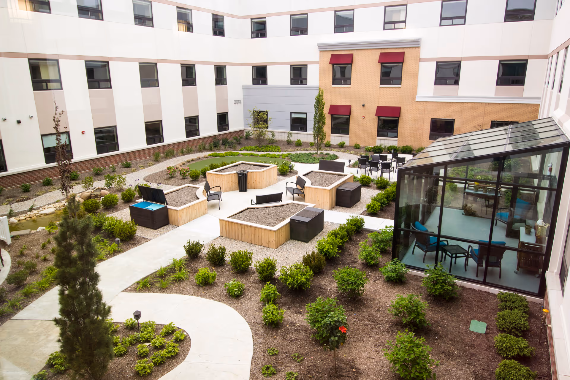 Outdoor courtyard area of The Avalon of Commerce Township featuring raised garden beds, seating areas with chairs and benches, a glass-enclosed sunroom with chairs and tables, surrounded by a multi-story building with windows and red awnings.
