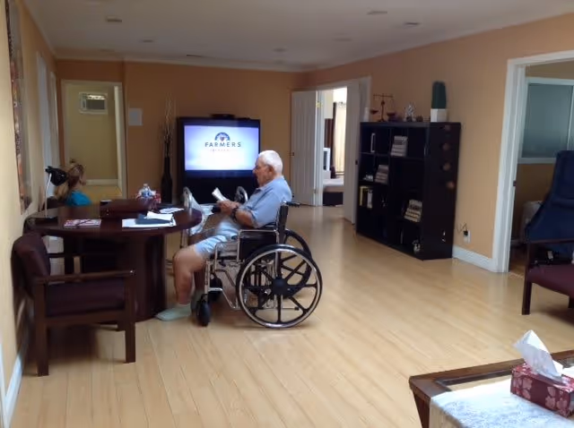 A spacious living room in a senior care facility with light wooden flooring and beige walls. An elderly man in a wheelchair is reading a book at a round wooden table, where a woman is also seated. A television is on in the background, and a black shelving unit with books and decorative items is against the wall. The room has multiple doorways leading to other areas.