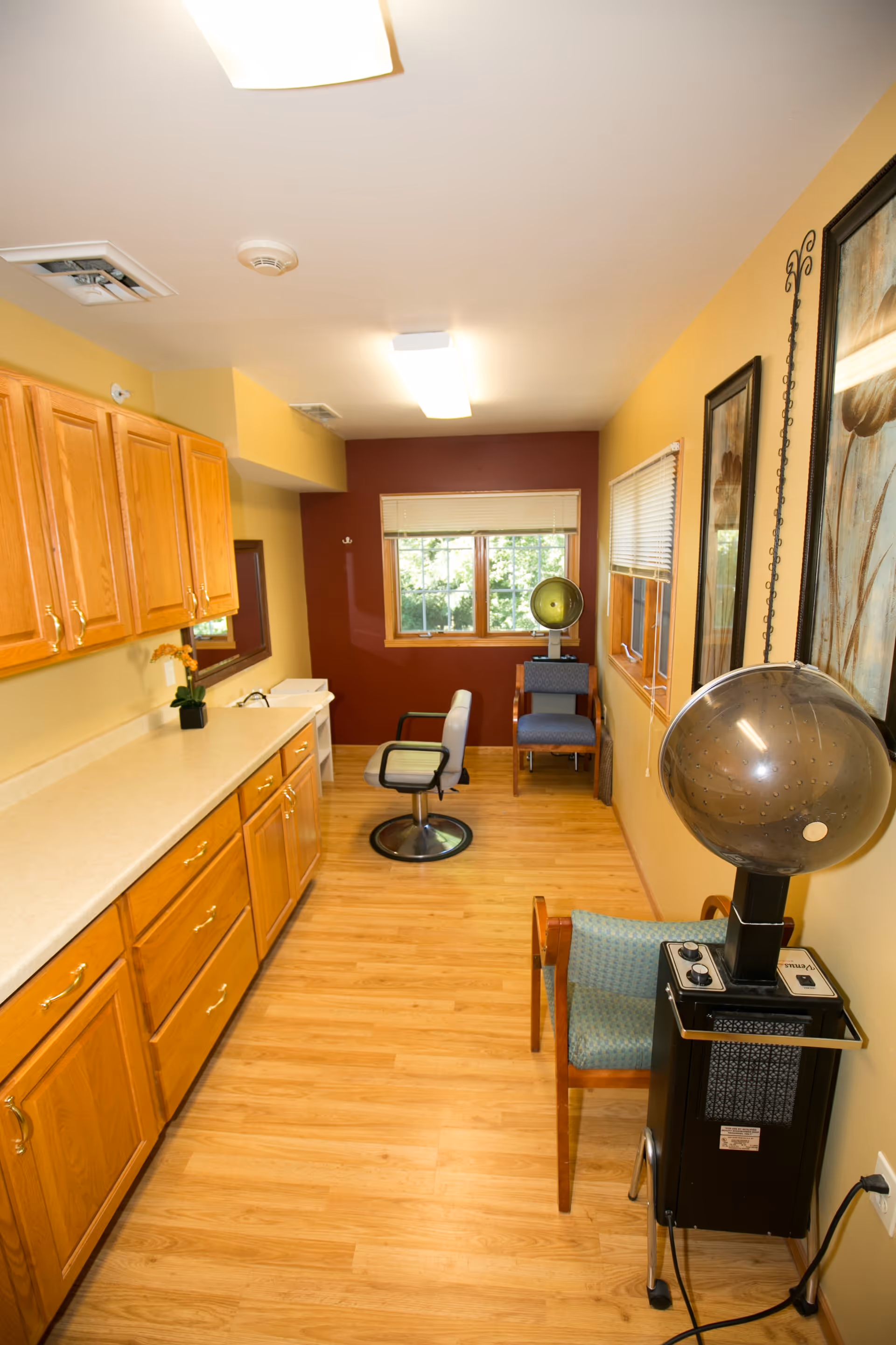 Interior room with wooden cabinets and countertop on the left, a salon chair in the center, two blue chairs with hair drying machines on the right, and a window at the far end with blinds partially open.