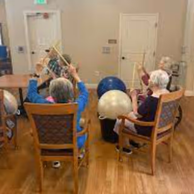 A group of elderly individuals seated in wooden chairs arranged in a circle in a room with wooden flooring. They are holding drumsticks and appear to be participating in a group drumming activity using large exercise balls placed in front of them.