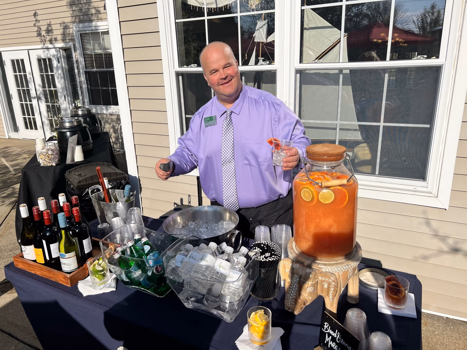 A man wearing a light purple shirt and checkered tie stands behind a table set up outdoors with various beverages including bottles of wine, sparkling water, bottled water, and a large glass dispenser filled with blood orange margarita garnished with citrus slices. The man is holding a drink with ice and a slice of blood orange, smiling at the camera. The table is covered with a dark blue cloth and is positioned in front of a beige building with white-framed windows.