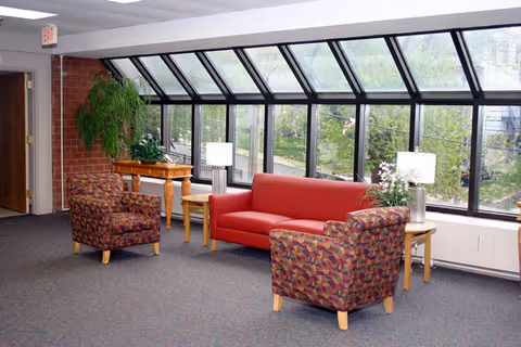 A bright indoor seating area with a red sofa and two patterned armchairs arranged around wooden side tables with lamps. Large slanted windows provide a view of greenery outside. There is a wooden table with plants and a brick wall on the left side.