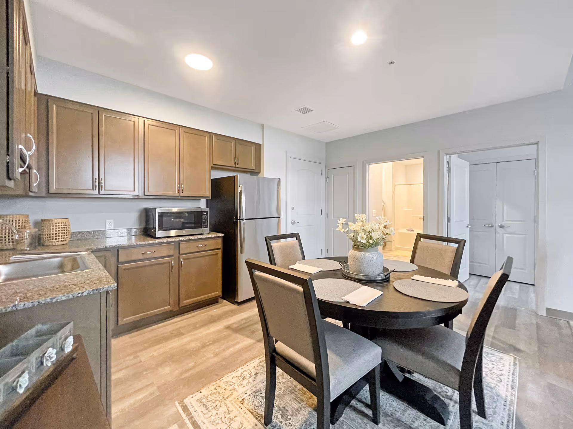 A modern kitchen and dining area featuring wooden cabinets, a stainless steel refrigerator, a microwave, and a round dining table set with four chairs and placemats. The room has light-colored walls and flooring, with a doorway leading to a bathroom and another room.