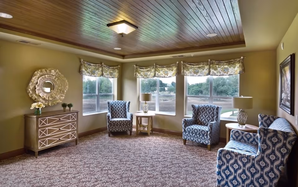 Sunlit common sitting room with patterned armchairs, side tables, a decorative dresser and large windows beneath a wood-paneled ceiling.