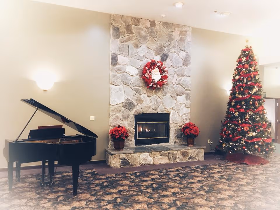 A cozy interior space featuring a stone fireplace with a red Christmas wreath above it, two red poinsettia plants on either side of the fireplace, a decorated Christmas tree with red ribbons and ornaments to the right, and a black grand piano to the left. The room has beige walls and patterned carpet flooring.