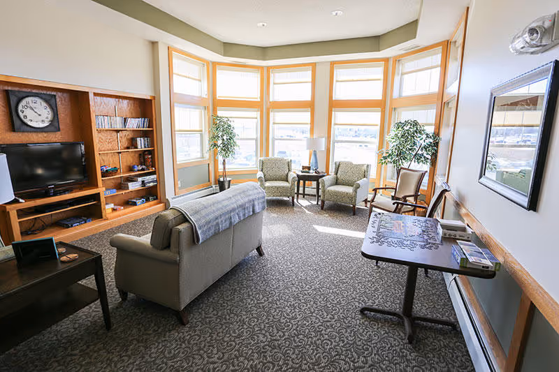 A bright living room with large bay windows letting in natural light. The room features a beige sofa with a gray throw blanket, two patterned armchairs, a wooden rocking chair, a small round table with a lamp, and a wooden entertainment center with a TV and shelves. There are two potted plants near the windows and a rectangular table with a puzzle on it against the right wall.