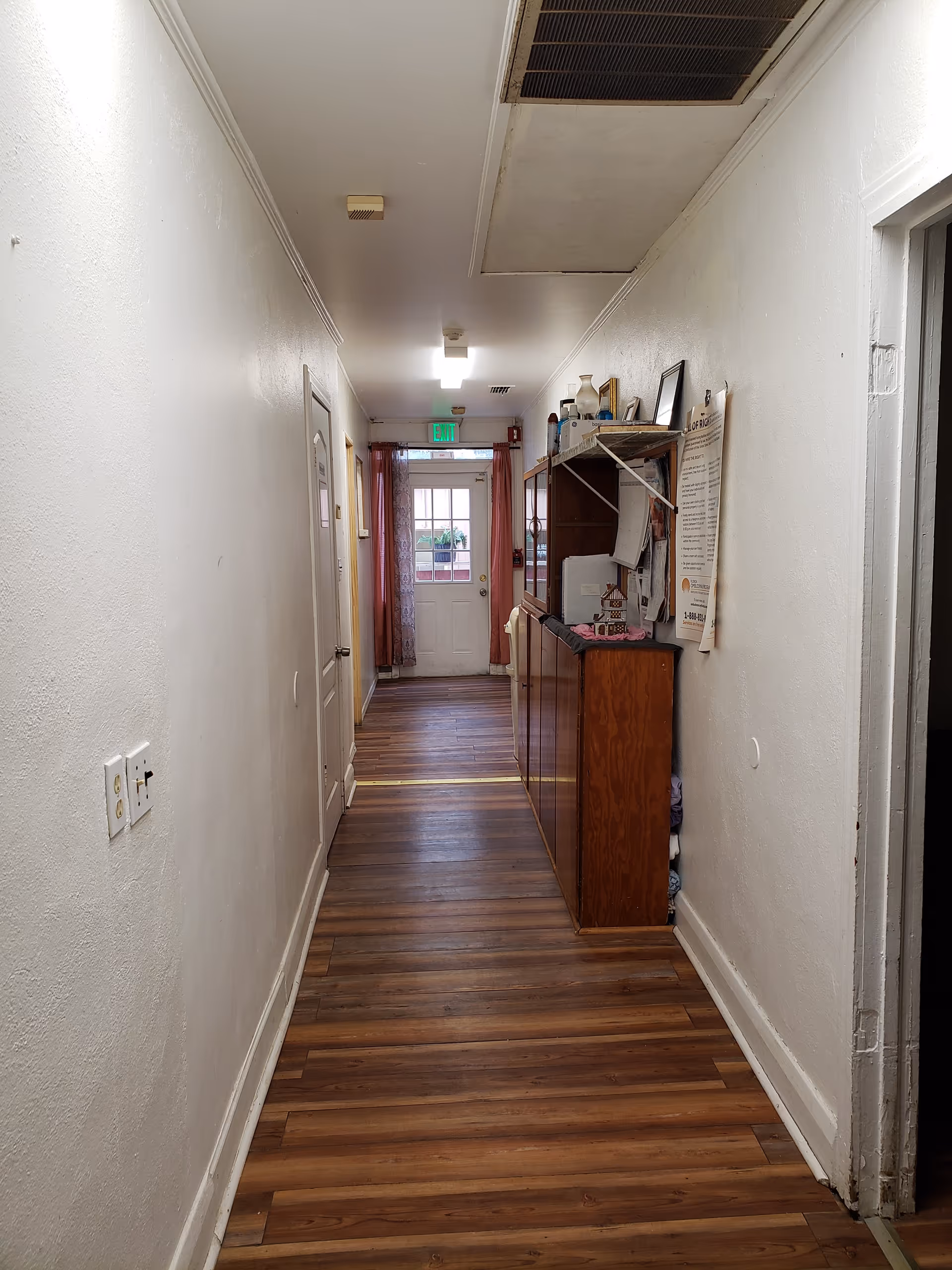 A narrow hallway with wooden flooring and white walls. On the right side, there is a wooden cabinet with various items on top and a shelf above it. At the end of the hallway, there is a door with a window and red curtains on either side. The ceiling has vents and lighting fixtures.