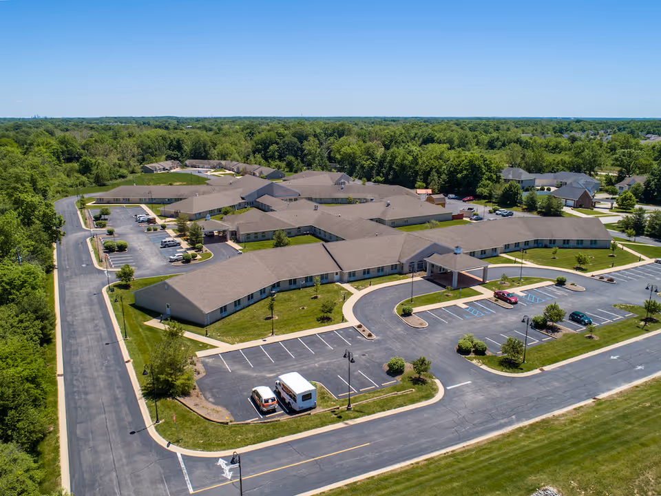 Aerial view of Avon Health and Rehabilitation Center, showing a large single-story building with multiple wings surrounded by parking lots and green lawns. The facility is located in a suburban area with trees and houses in the background under a clear blue sky.