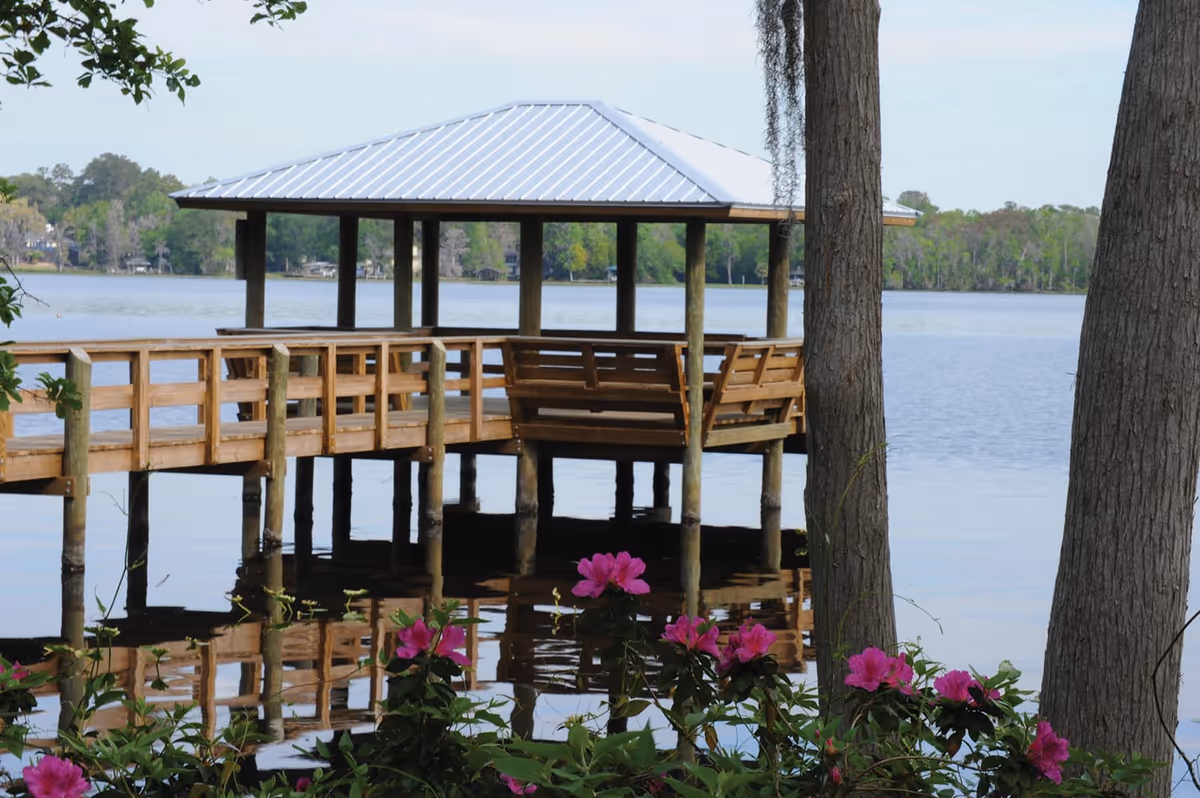 A wooden gazebo with a metal roof extends over a calm lake, supported by wooden pillars. The gazebo has built-in wooden benches and is connected to the shore by a wooden walkway. In the foreground, pink flowers and green foliage frame the scene, with two tree trunks on the right side. The background shows a tree-lined shore across the lake under a clear sky.