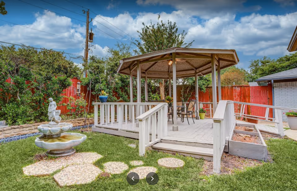 Outdoor garden area with a white wooden gazebo featuring a table and chairs underneath. There is a small stone fountain with a statue in the foreground, surrounded by green grass and stepping stones leading to the gazebo. The area is enclosed by a red wooden fence and various bushes and trees. The sky is partly cloudy.