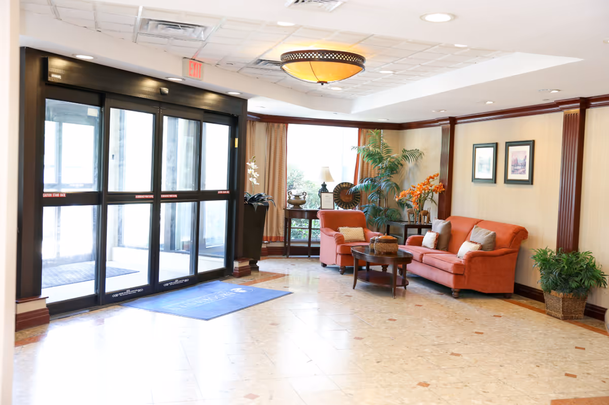 A bright and welcoming lobby area with automatic glass sliding doors on the left and a seating area on the right featuring an orange sofa, matching armchair, a wooden coffee table, potted plants, framed artwork on the wall, and a ceiling light fixture.