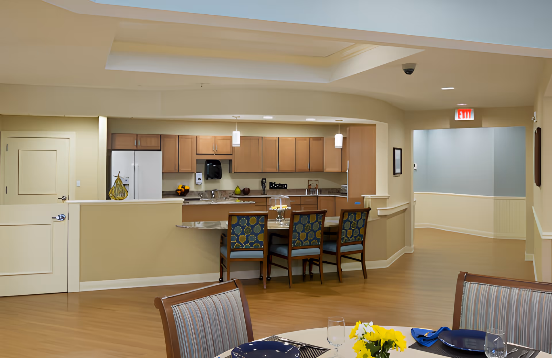 Interior view of a senior living facility kitchen and dining area with wooden cabinets, a refrigerator, a countertop with three chairs, and a dining table set with plates, glasses, and a yellow flower centerpiece. The space has light-colored walls and wood flooring, with an exit sign visible in the hallway.