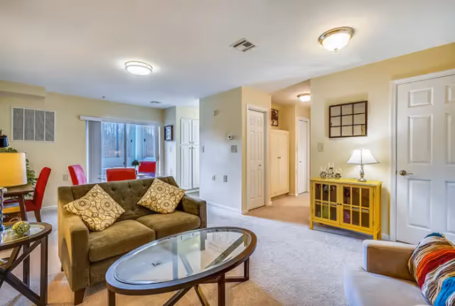 A cozy living room area in a senior living facility with a brown sofa adorned with patterned cushions, a glass coffee table, a yellow cabinet with a lamp on top, and a hallway leading to other rooms. The room is well-lit with ceiling lights and has beige walls and carpeted floors.