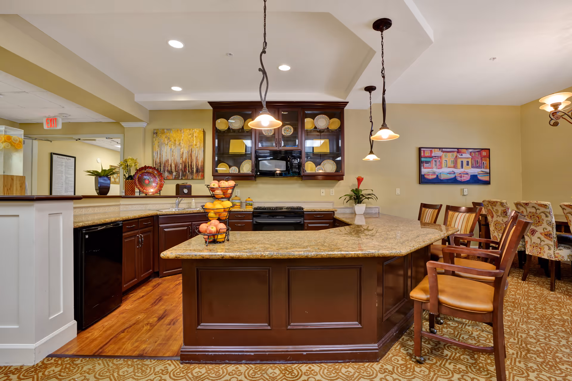 A spacious kitchen area with a large granite countertop island surrounded by wooden chairs with cushioned seats. The kitchen features dark wood cabinets, a black stove, microwave, and dishwasher. Decorative plates are displayed in glass-front cabinets above the stove. The room is well-lit with recessed ceiling lights and hanging pendant lights. There are colorful paintings on the walls and a carpeted floor with a patterned design.