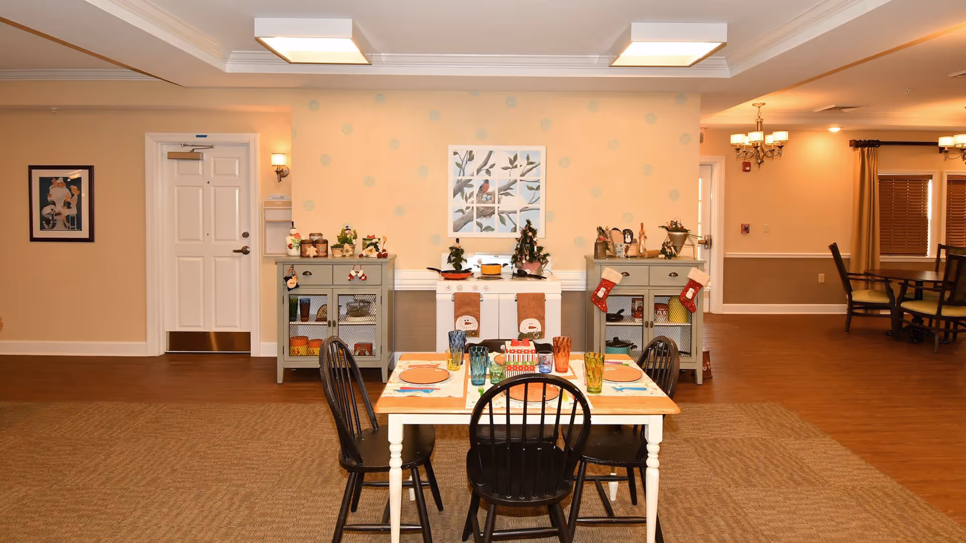 A cozy dining area in a senior living facility with a wooden table set for four, featuring colorful glasses and plates. Behind the table are two small cabinets decorated with holiday stockings and festive ornaments, and a white play stove with small Christmas trees on top. The walls are painted beige with a framed bird artwork in the center. The room has warm lighting and wooden flooring.
