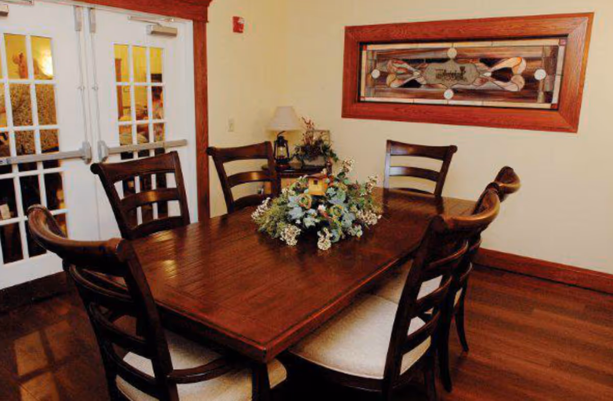 Dining room with a wooden rectangular table, six chairs and a floral centerpiece in front of French doors and a stained-glass window.