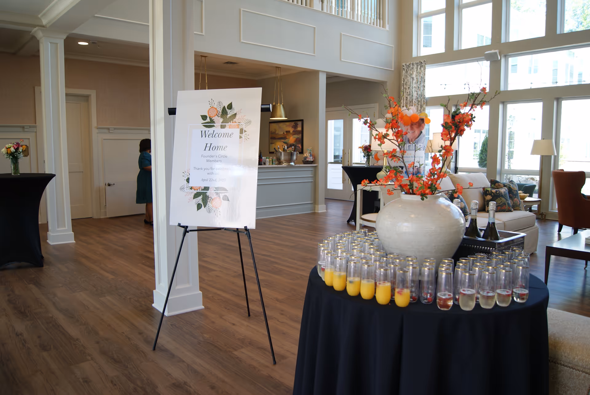 A bright and spacious interior room with large windows letting in natural light. In the foreground, a round table covered with a black tablecloth holds many glasses filled with orange juice and champagne, arranged around a large white vase with orange flowers. To the left, a sign on an easel reads 'Welcome Home Founder's Circle Members Thank you for celebrating with us! April 22nd, 2023'. The room has wood flooring, white pillars, and comfortable seating areas with sofas and chairs in the background.