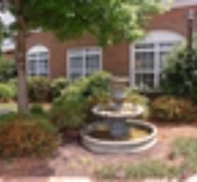 Outdoor garden area with a tiered water fountain surrounded by bushes and trees, in front of a brick building with white-framed windows.