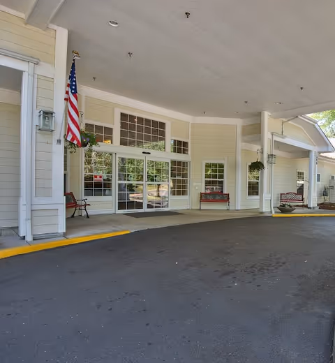 Covered front entrance and drive-up drop-off of a light-colored assisted living building with an American flag and benches.