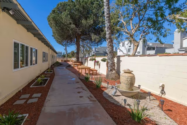 Outdoor walkway at Victoria Healthcare and Rehabilitation with a paved path, benches, a large decorative urn fountain, plants, and trees along the sides, bordered by a beige building on the left and a white wall on the right under a clear blue sky.