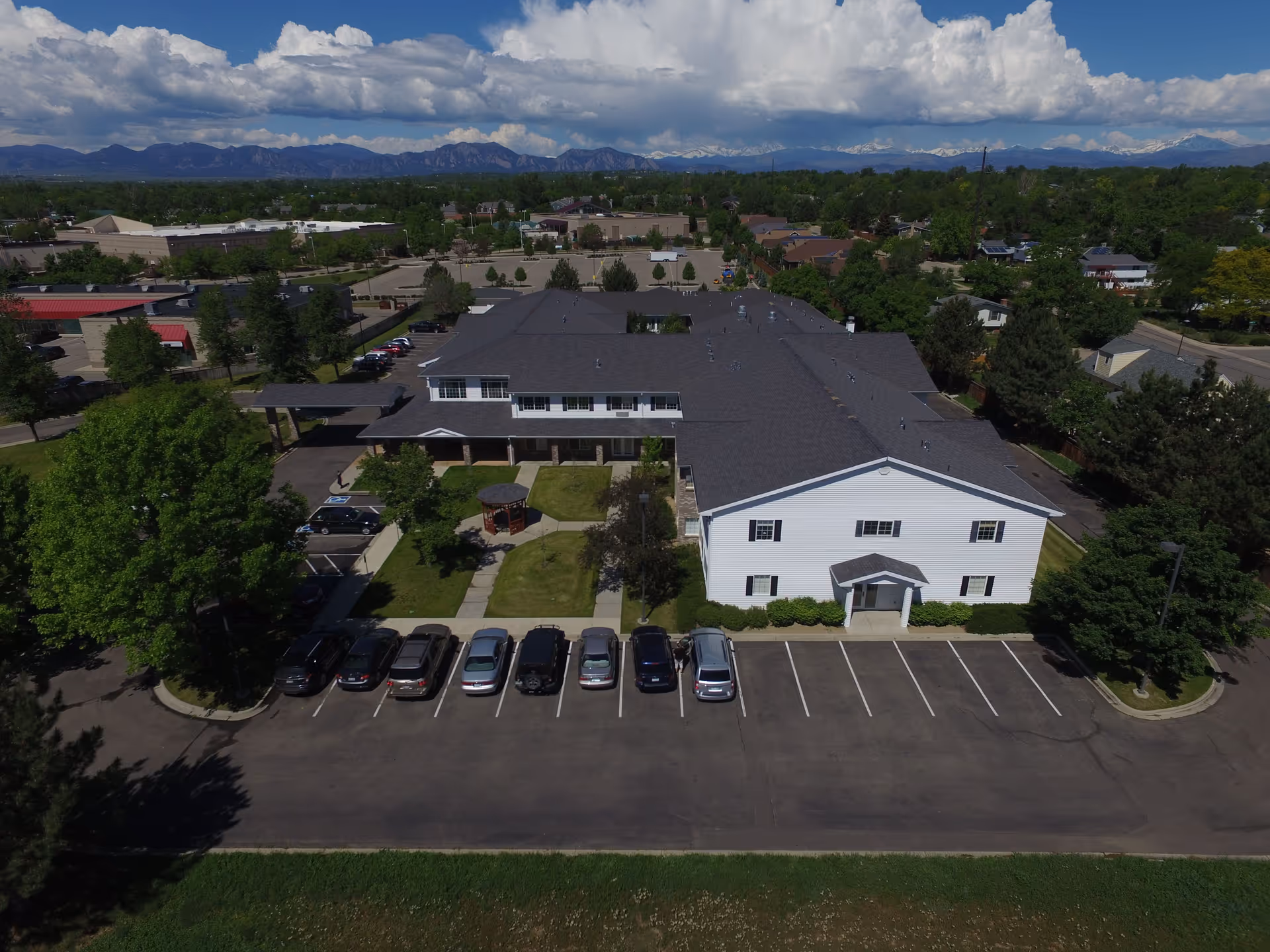 Aerial view of Waneka Park Assisted Living facility showing a large two-story building with a dark roof and white exterior walls. There is a parking lot with several cars in front of the building, green lawns, trees, and a small gazebo in the courtyard. In the background, there are more buildings, trees, and a mountain range under a partly cloudy sky.