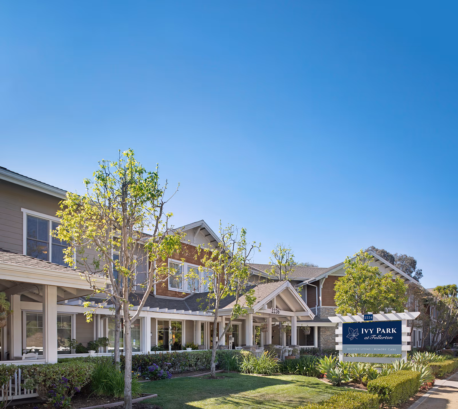 Exterior view of Ivy Park at Fullerton, a senior living facility, showing a two-story building with beige and brown siding, a covered entrance, landscaped greenery, and a clear blue sky.