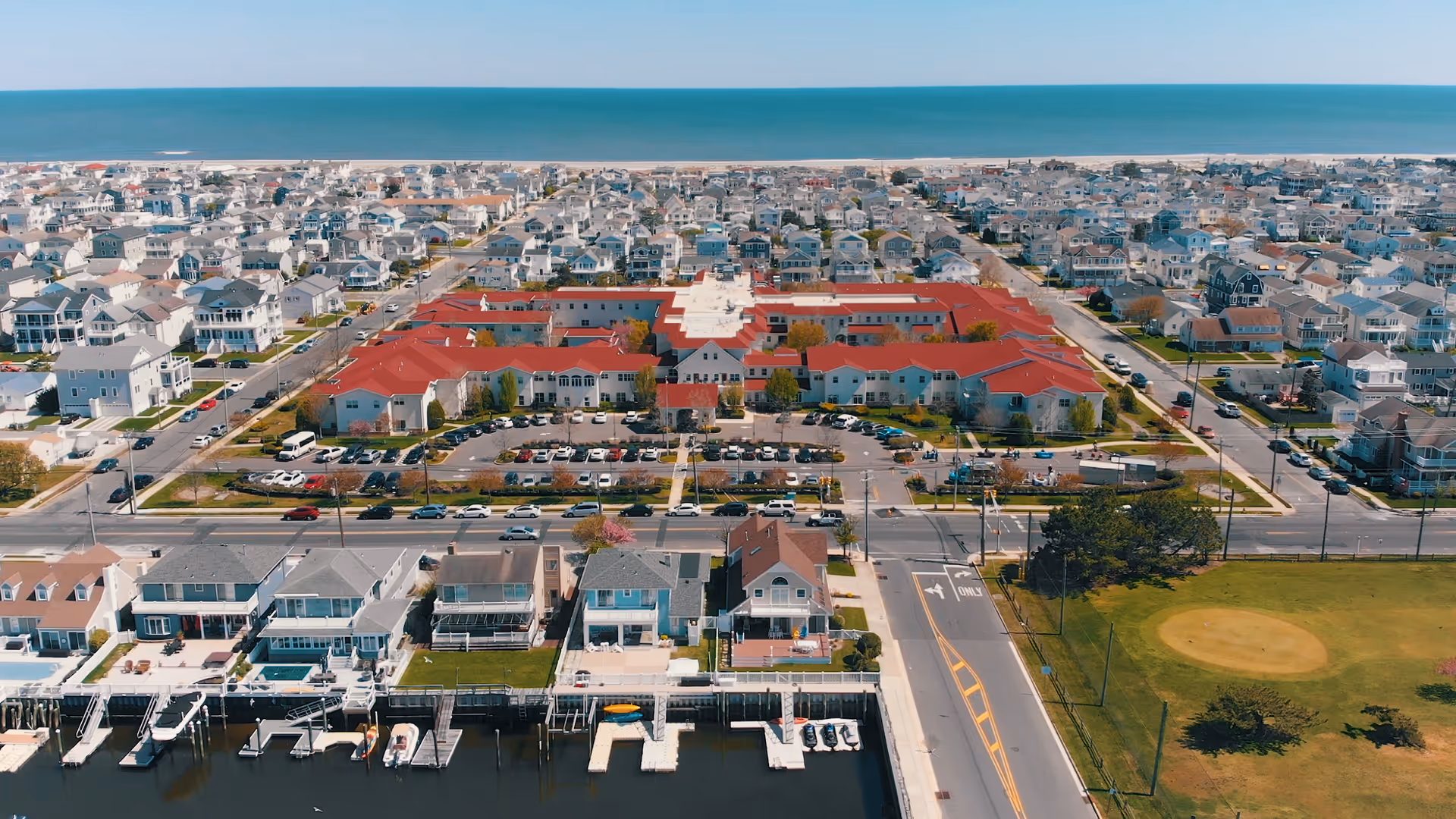 Aerial view of United Methodist Communities at The Shores, a large residential facility with red roofs, surrounded by a neighborhood of houses near the ocean. There is a parking lot in front of the building and a small marina with boats docked along the waterfront.