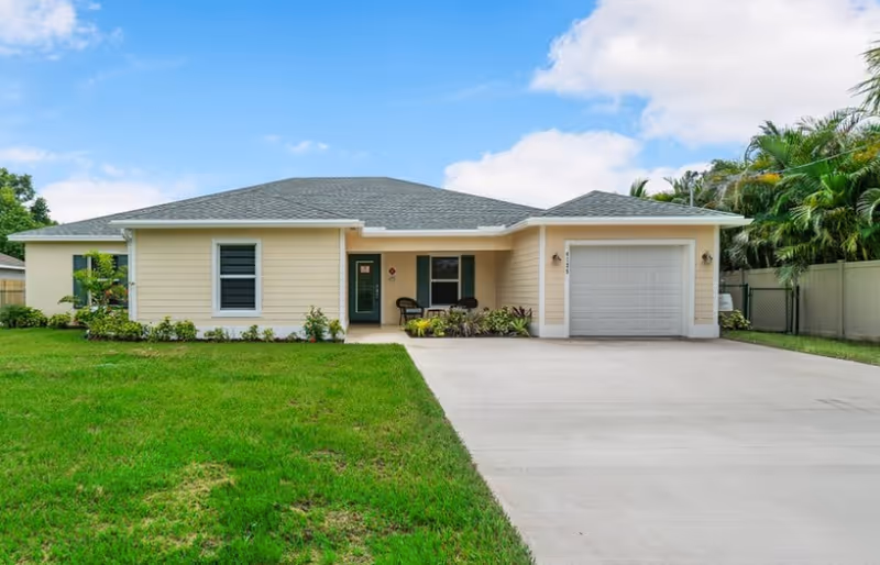 Single-story house with light yellow siding, a gray shingled roof, a front porch with two chairs and plants, a single garage door, and a concrete driveway surrounded by a green lawn and palm trees under a partly cloudy sky.