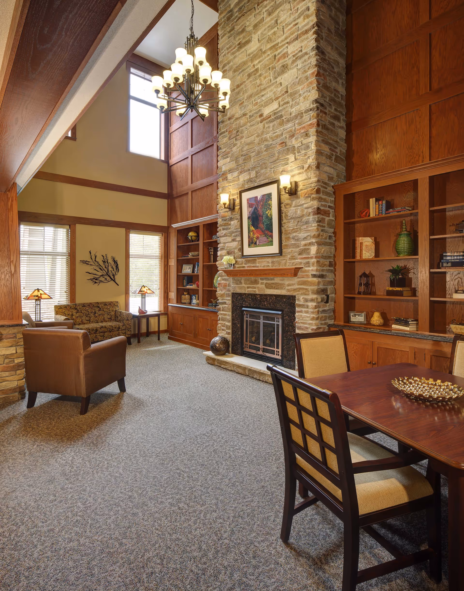 A cozy living room area in an assisted living facility featuring a tall stone fireplace with a framed picture above it, built-in wooden bookshelves on either side, a brown leather armchair, a patterned sofa, two table lamps, and a wooden dining table with cushioned chairs. The room has large windows allowing natural light and a chandelier hanging from the high ceiling.