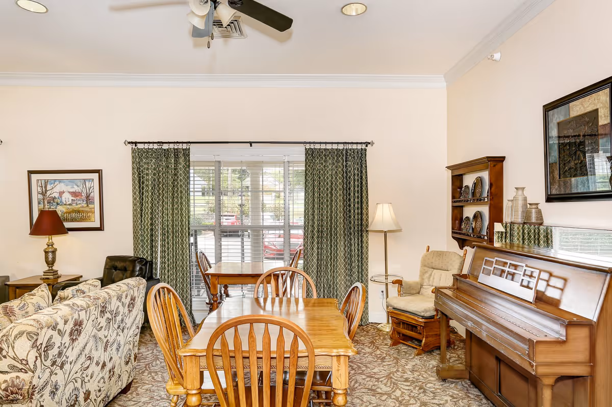 A cozy living room area with a floral patterned sofa, wooden dining table with chairs, a wooden piano, a cushioned rocking chair, a floor lamp, and framed artwork on the walls. Green patterned curtains cover a large window with blinds, and a ceiling fan is visible above.
