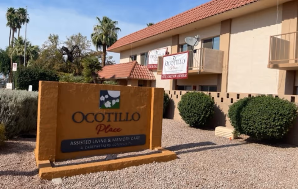 Exterior view of Ocotillo Place assisted living and memory care facility with a large sign in the foreground and a two-story building with balconies and a tiled roof in the background. There are bushes and palm trees around the property under a clear sky.