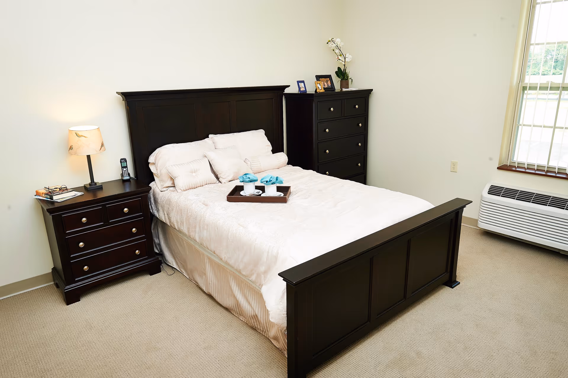 Neatly made bed with a dark wooden headboard and footboard in a bright bedroom with a matching nightstand and dresser, lamp, tray on the bed, and a window air unit.