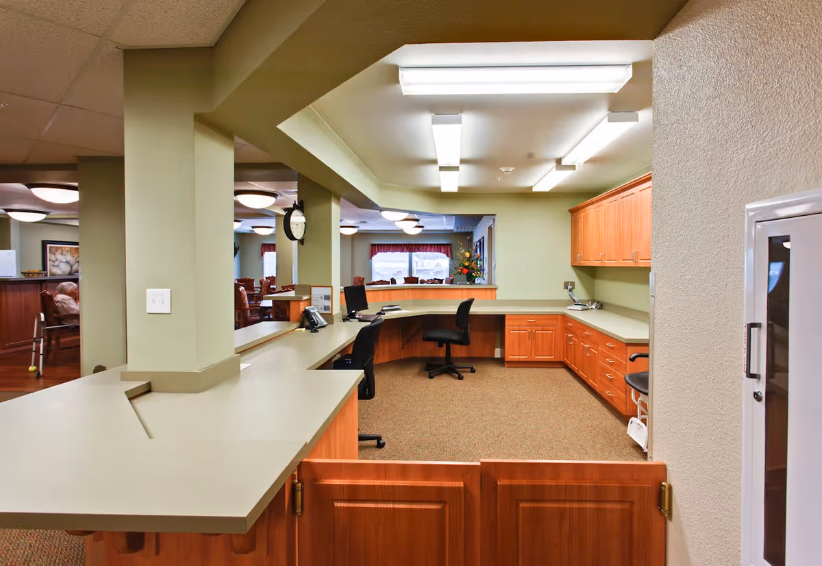 Interior view of a senior living community office or reception area with a large L-shaped desk, office chairs, wooden cabinets, and overhead fluorescent lighting. In the background, there is a dining area with tables and chairs, and a person sitting with a walker nearby.
