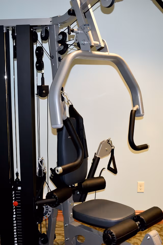 A multi-function gym machine with padded seat and backrest, handles, and weight stack, set against a plain wall with an electrical outlet visible.