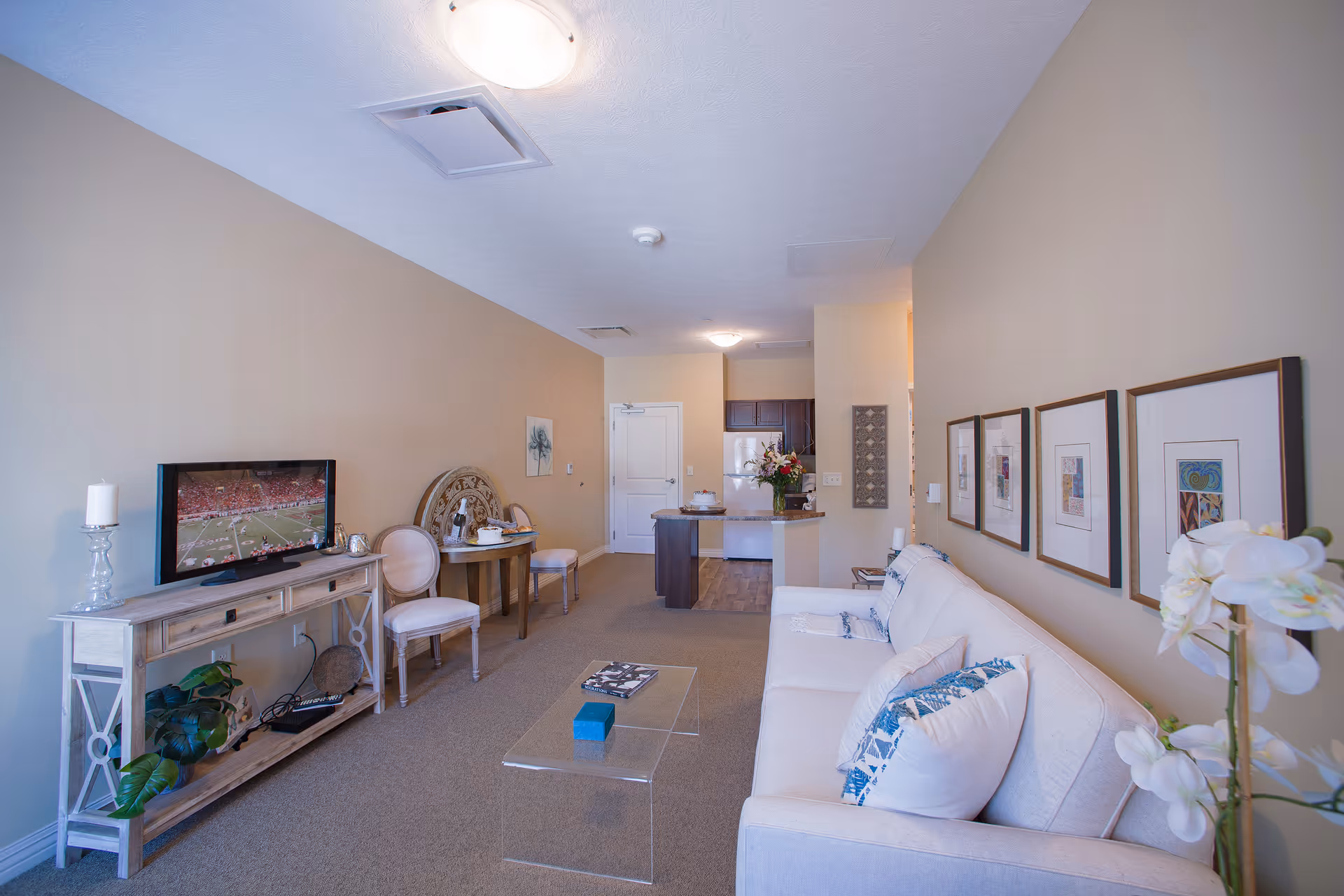 A cozy senior living room with a white sofa adorned with blue and white pillows, a clear glass coffee table, a wooden TV stand with a television showing a football game, two chairs and a small round table, and a kitchen area in the background. The walls are beige with framed artwork and a white orchid plant is visible in the foreground.