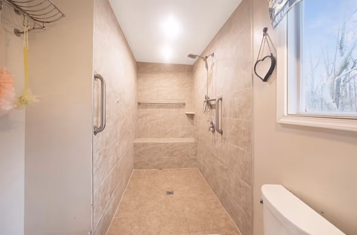 A spacious walk-in shower with beige tiled walls and floor, equipped with multiple grab bars and a built-in bench. There is a handheld showerhead attached to the wall, and a window with a heart-shaped hanging decoration is visible on the right side. Part of a white toilet is also visible in the lower right corner.
