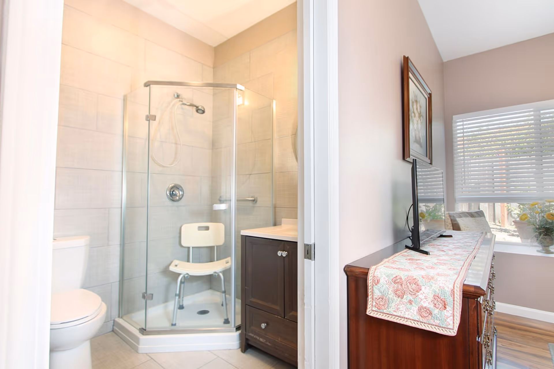 A bathroom with a glass-enclosed corner shower featuring a white shower chair, a toilet, and a dark wood vanity with a sink. Adjacent to the bathroom is a room with a wooden dresser topped with a floral runner, a flat-screen TV, a framed picture on the wall, and a window with white blinds letting in natural light.