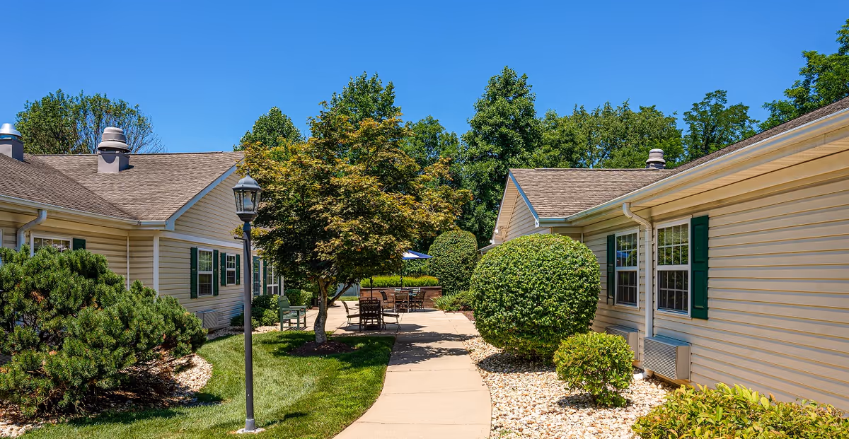 Outdoor view of a senior living facility courtyard with a paved walkway, manicured bushes, trees, and outdoor seating with tables and chairs between two beige buildings under a clear blue sky.