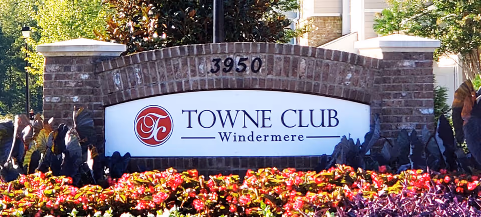 Brick sign with the address number 3950 and a white panel displaying the Towne Club Windermere logo and name, surrounded by colorful flowers and greenery.