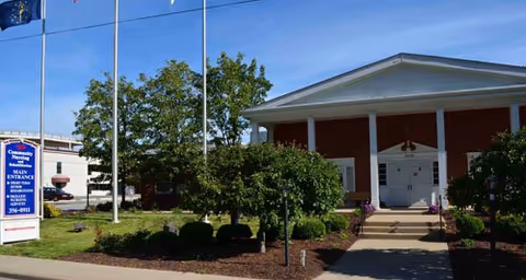 Front exterior view of Community Nursing and Rehabilitation facility with a white entrance building, steps leading up to the door, landscaped bushes and trees, and a sign indicating the main entrance and contact information.