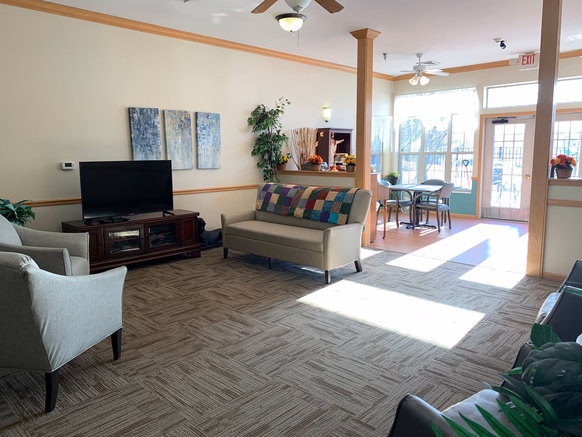 A bright and cozy living room area with a beige sofa adorned with a colorful patchwork quilt, two gray armchairs, a flat-screen TV on a wooden stand, and a small dining table with chairs near large windows letting in natural light. The room features light-colored walls with wooden trim and ceiling fans.