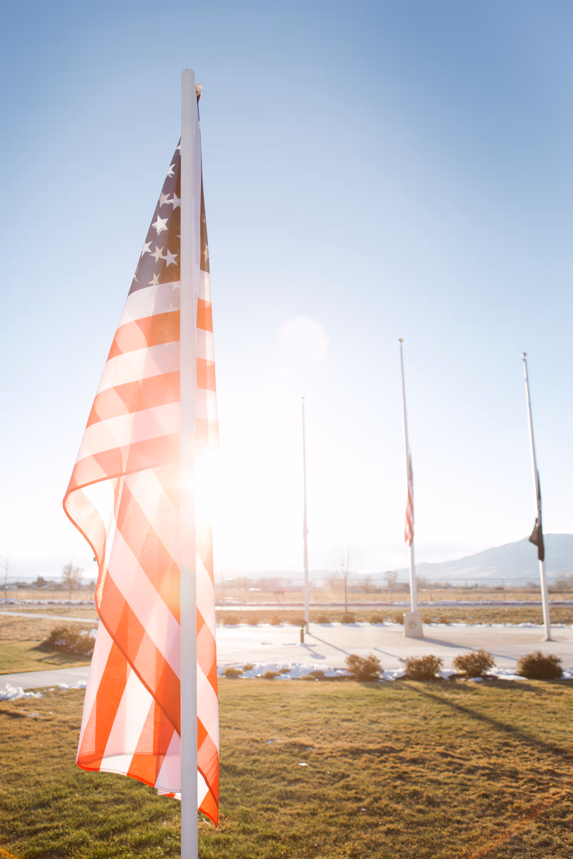 American flags flying at half-staff on flagpoles in a grassy outdoor area with a clear blue sky and mountains in the background, with sunlight shining through one of the flags.