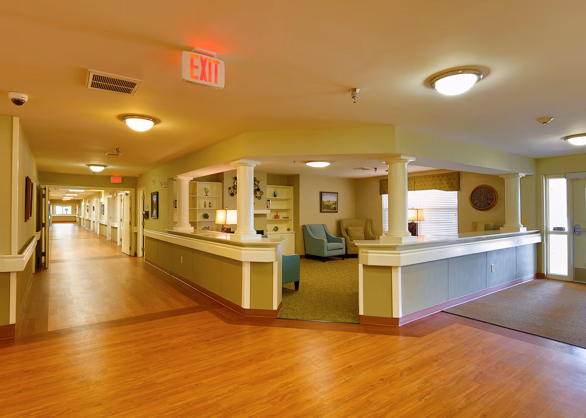 Interior view of a senior living facility hallway with wooden flooring and beige walls. To the right, there is a common sitting area with armchairs, lamps, and decorative shelves. The hallway extends into the distance with multiple doors and exit signs visible on the ceiling.
