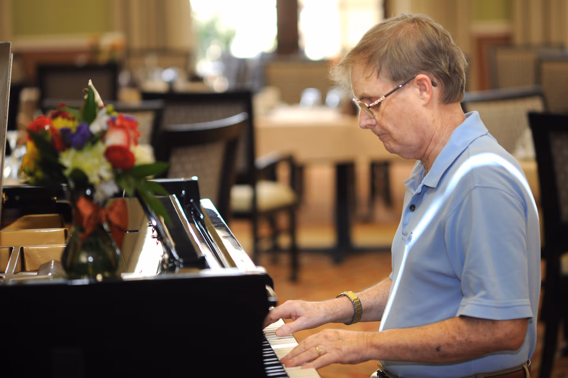 An elderly man wearing glasses and a light blue polo shirt is playing a black grand piano indoors. A vase with colorful flowers is placed on the piano. The background shows a dining area with tables and chairs, softly lit by natural light from windows.